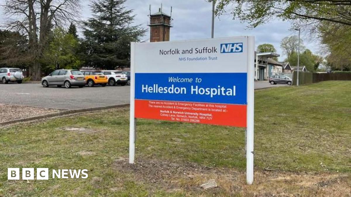 The picture is a close up of the sign for Hellesdon Hospital. It says Norfolk of and Suffolk NHS Foundation Trust  is white red and blue. It also says "Welcome to Hellesdon Hospital". There is a car park behind the sign and some of the cars are visible. The sign it is positioned on grass.