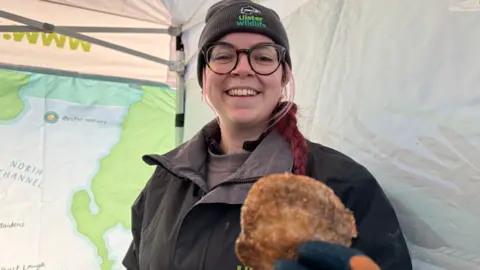 A woman, wearing a black cap, black rimmed glasses, and a black and grey rain jacket. Her hair, dyed red, is tied up in a baid. She is holding an oyster in her hand and smiling at the camera. She is standing instead a white tent with a map on the wall in the background.