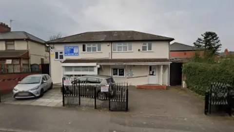 Google General view of the entrance of Naseby Medical Centre in Birmingham. A cream coloured building with a small driveway outside. Two cars are parked in the driveway which has some iron gates across part of it.