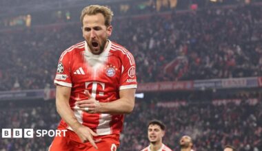 Harry Kane celebrates a goal for Bayern Munich, with his mouth wide open. He is wearing Bayern's largely red home kit, with white stripes on the sleeves, and a white collar and cuffs, with large crowd visible in the background