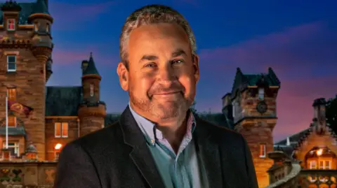 BBC/PA Handout photo of Hugo, one of the contestants in series four of BBC1's The Traitors. He wears a blue shirt, a navy blazer and the castle can be seen in the background behind him. 