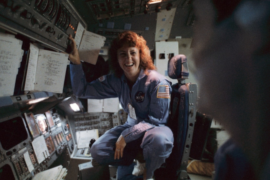 A woman in blue overalls crouches on a chair in a cockpit, grinning towards the camera.