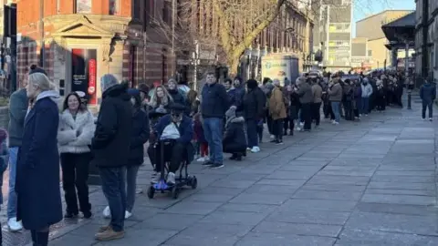 BBC A queue of people down a street in Preston. Many are wearing coats and bracing from the cold. 