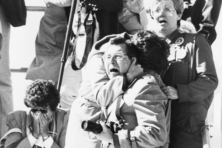 Black and white photo of people looking shocked, two embracing and others with hands over their faces.