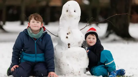 PA Media Two young boys dressed in warm weather gear are sitting on snowy ground on either side of a snowman they have built 