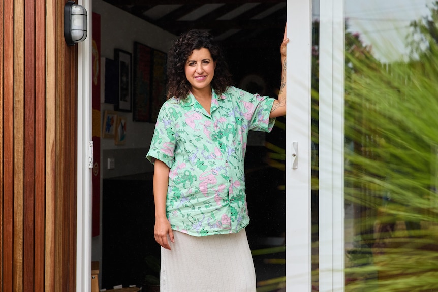 Yasmine beams while leaning against a doorframe in a patterned shirt while pregnant.