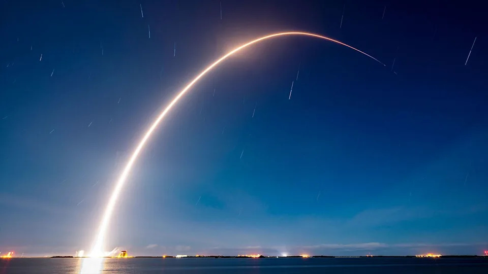 A time lapse photo captures the break streak of a rocket launching into the early morning dark blue sky over a body of water.