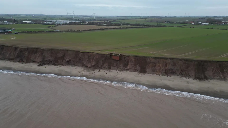 A wide shot of the eroded cliffs with waves breaking in front of them. The brick-built bunker appears to be unsupported on the cliff face. 
