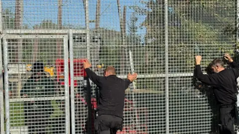 Lincolnshire Wildlife Park A tall metal gate with square holes in it with two men dressed all in back leaning their hands against it.