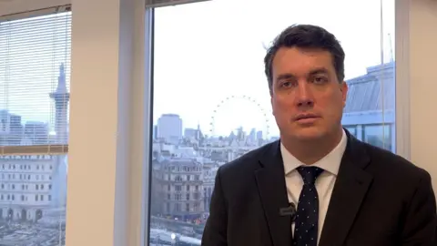 Jason Williams, wearing a dark suit, stands in the offices of the SFO, overlooking Trafalgar Square, with the London Eye in the distance.