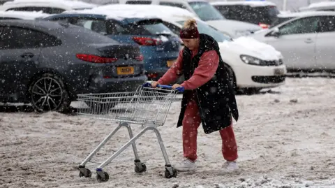 Reuters A woman pushes a trolley full of her supermarket shopping through far-stretching, thick brown slush in a car park. She wears a black sleeveless coat and red tracksuit. Her trolley is empty. In the background snow covered cars are visible 