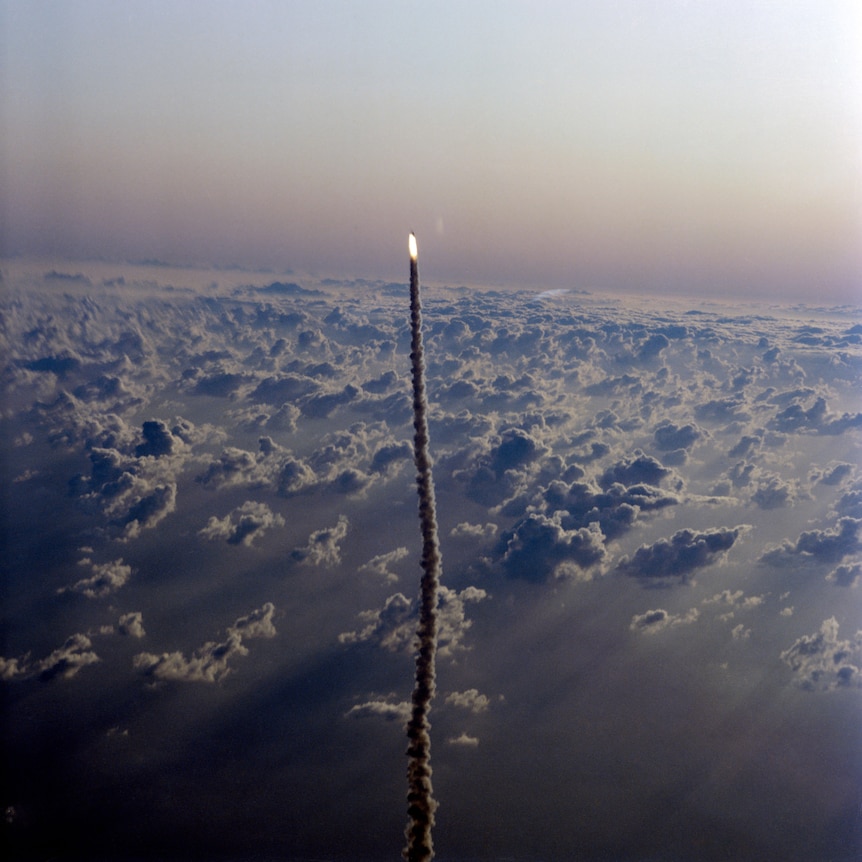 Distant aerial shot of a rocket trailing a pillar of smoke above billowing clouds.