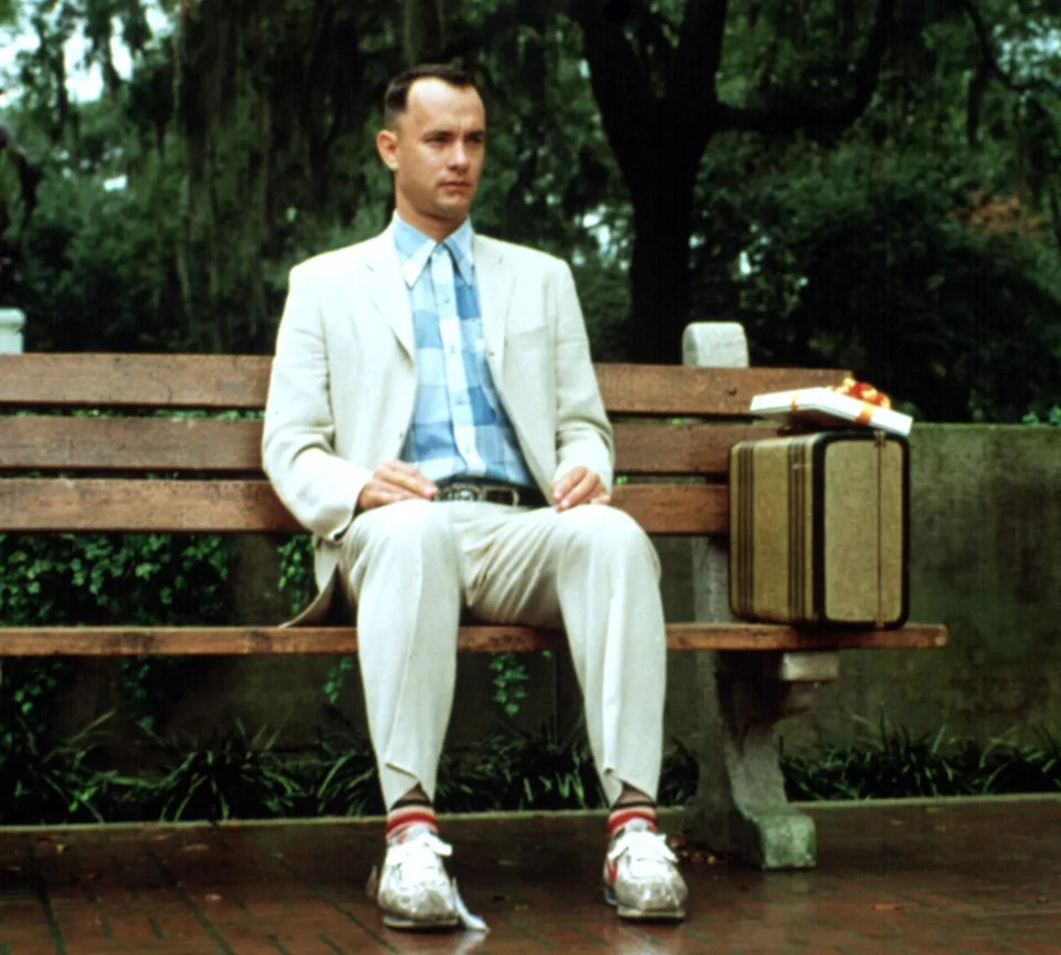 Tom Hanks sitting on a bench with a suitcase and box of chocolates beside him in "Forrest Gump"