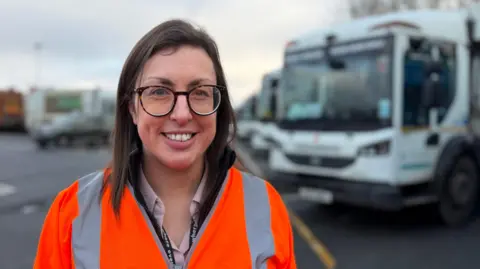Becki Bowden / BBC A woman with shoulder-length brown hair and glasses smiles as she stands in front of a row of bin lorries, which are out of focus. She is wearing a hi-vis orange jacket.