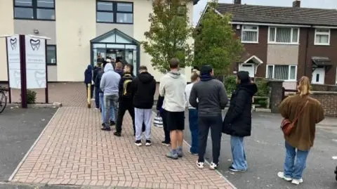 BBC A queue of dozens of people is formed outside a dentist's practice. 