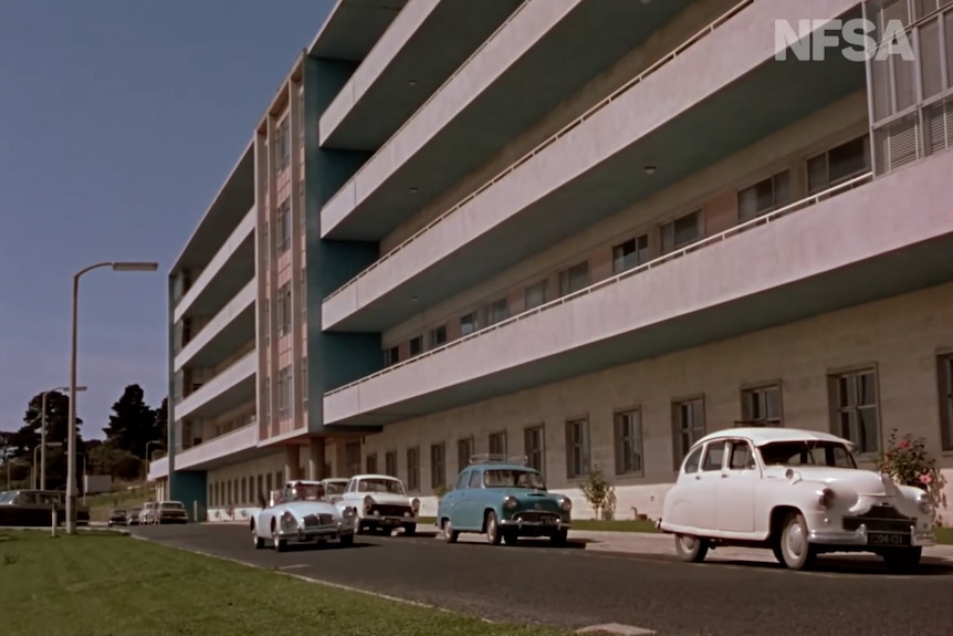 An old restored photo of a wide white building with many stories and old cars parked outside alongside the ground floor.