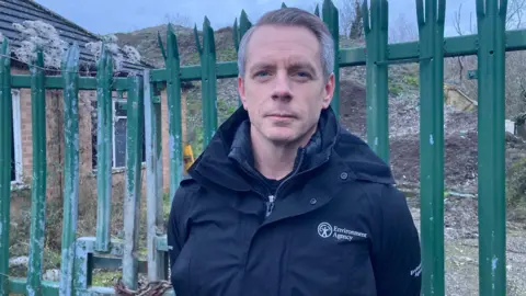 BBC A man wearing an Environment Agency jacket, standing in front of a green bolted gate - behind that is a pile of waste that was dumped there illegally.