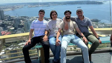 Ekam Dhaliwal A family of four are in a tall building with glass overlooking a city and the sea. There are two young men - the sons, and a mother and father. They all have dark hair and are sitting close together, smiling