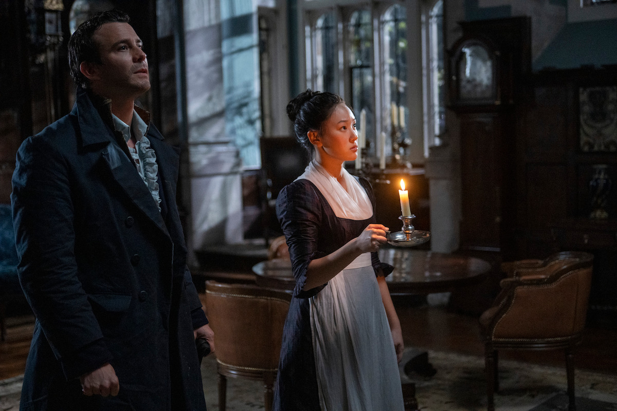 A man and woman in period clothing stand in a dimly lit, elegant room. The woman holds a lit candle, illuminating their faces, while antique furniture and large windows are visible in the background.