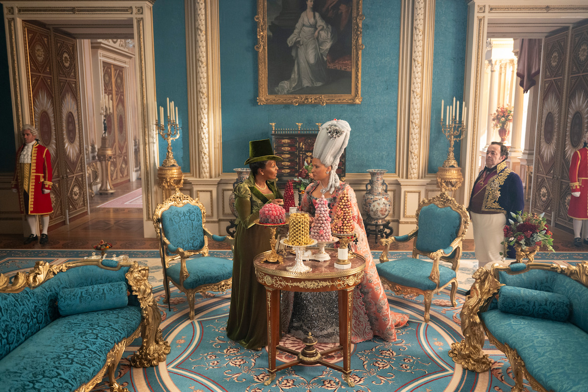 Two women in elaborate, historical costumes stand by a table with desserts in a lavish, ornate blue and gold palace room, surrounded by elegant furniture, chandeliers, guards, and a portrait on the wall.