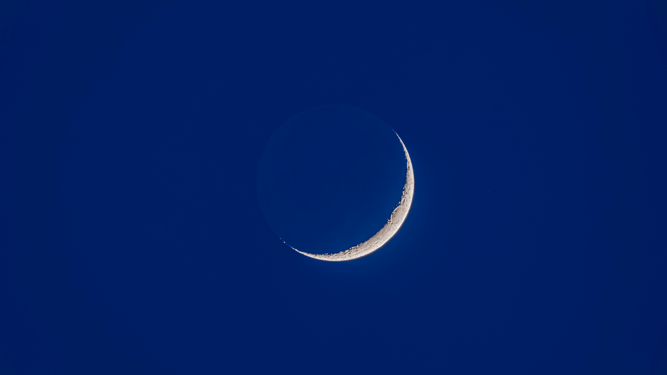 A thin crescent moon is photographed in a deep blue evening sky. It's unlit side can be seen shining subtly with light that has reflected off Earth's surface.