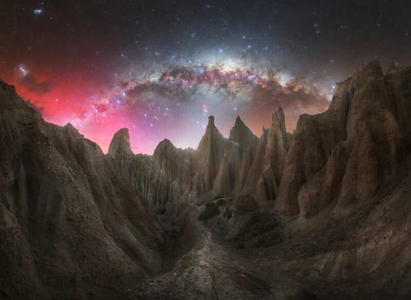 Jagged rock formations rise dramatically beneath a vibrant night sky, illuminated by a sweeping arc of the Milky Way and a red and purple glow near the horizon. The scene is otherworldly and awe-inspiring.