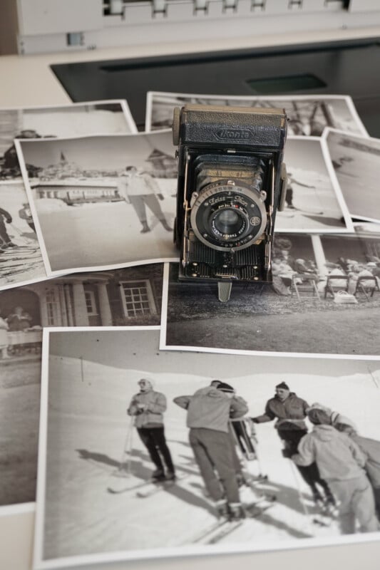 An old folding camera is placed on top of several black-and-white vintage photographs, some showing people skiing and others featuring groups near buildings. The background includes a modern office device.