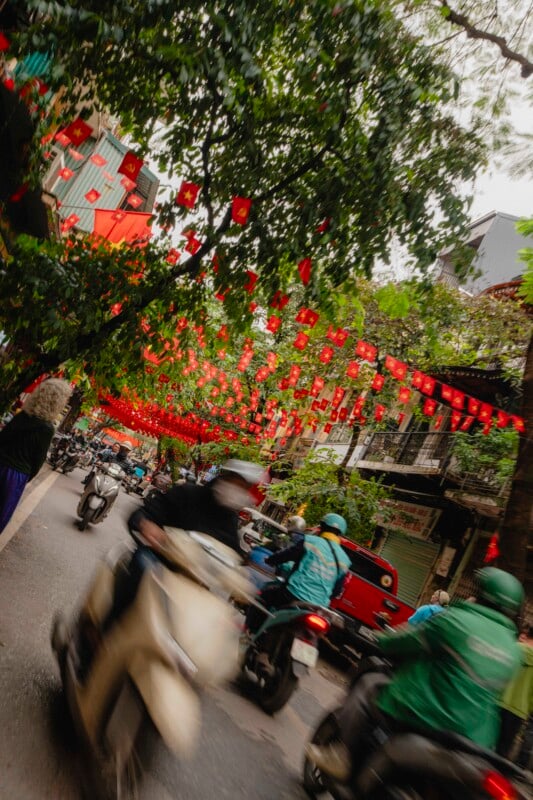 A busy street scene with blurred motorbikes in motion, trees overhead, and many red lanterns hanging across the road. Buildings with balconies line the street, creating a lively, festive atmosphere.
