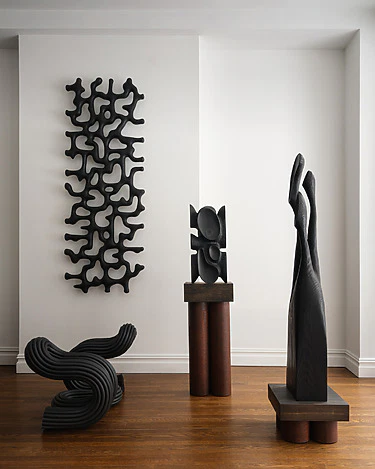 Modern abstract sculptures on a wooden floor with white walls, featuring geometric and organic shapes in dark tones.