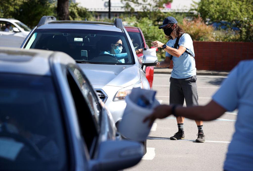 Photo looking head-on at a line of stopped cars. The driver in the second car receives a bucket from a person standing outside the car. Both people are wearing masks over their mouths.