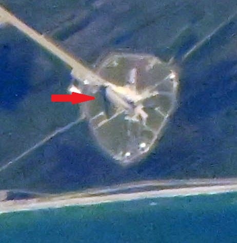 Aerial view of a launch pad complex near a shoreline, featuring circular and linear structures. A red arrow points to a specific area on the pad.