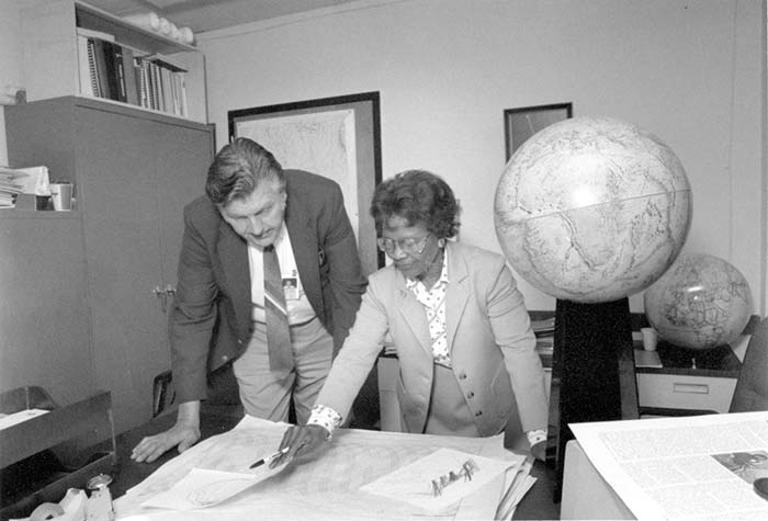 A man and a woman stand in an office, looking at maps spread out on a desk. A large globe is visible in the background.