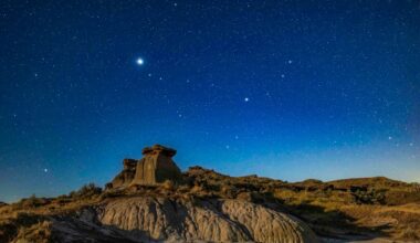 A glowing bright blue night sky full of white stars looks over a rocky landscape.