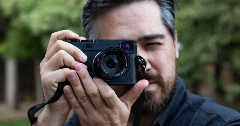 A man with a beard is holding a black camera up to his face, looking through the viewfinder and preparing to take a photo. The background is blurred greenery.