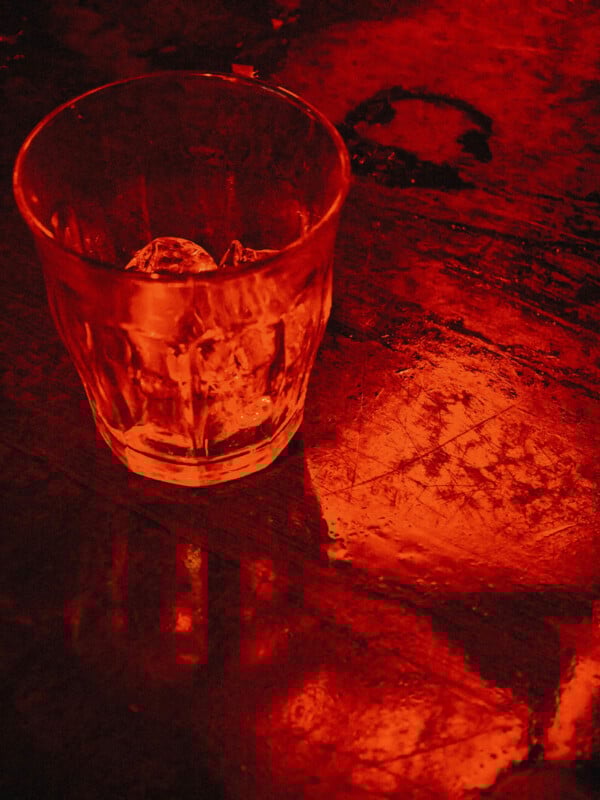 A glass with ice cubes sits on a scratched, reflective surface, illuminated by intense red lighting, creating dramatic shadows and highlights.