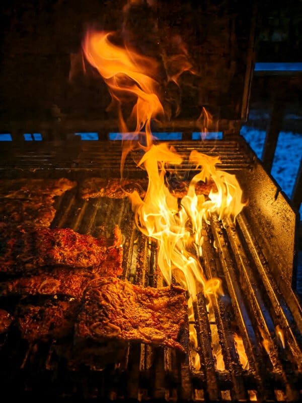 Flames rise on a grill as several pieces of meat cook, with a bright fire on the right side and charred grill marks visible on the meat and grates.