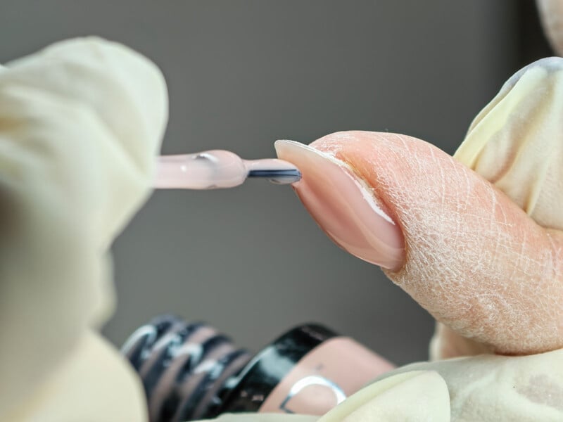 A gloved hand applies a coat of light pink nail polish to a fingernail with a brush, focusing closely on the nail and the precision of the manicure process.