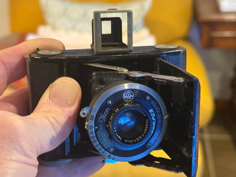 A hand holding a vintage folding camera with a round lens and metal dials. The camera’s viewfinder is popped up, and the background is softly blurred with a yellow chair and a wooden table visible.