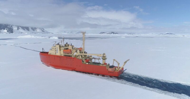 A large red icebreaker ship cuts through thick sea ice in a polar region, with snow-covered mountains and a cloudy sky in the background.