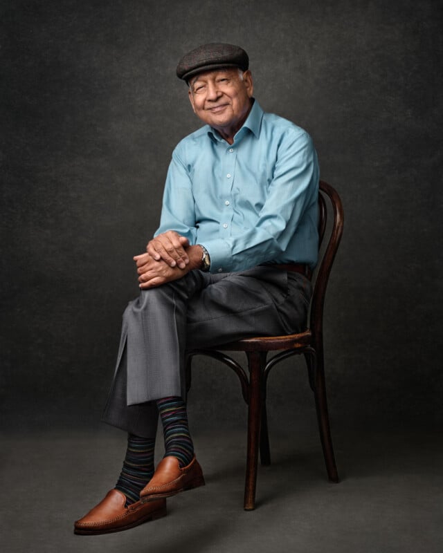 An older man wearing a flat cap, light blue shirt, gray trousers, and brown shoes sits on a wooden chair, smiling with his hands folded over his knee in front of a dark, textured background.