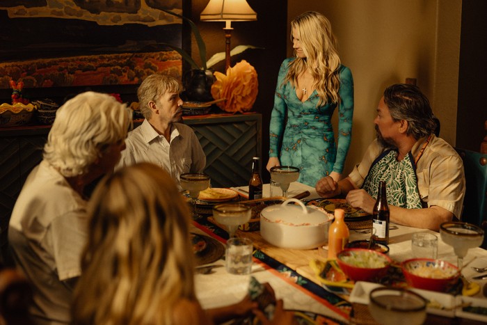 A group of adults sit around a dining table in a warmly lit interior, mid-meal. A woman in a fitted teal dress stands at the table, drawing the attention of the others as they look up at her. Plates of food, drinks, and serving dishes are spread across the table, suggesting a shared dinner. The room is richly decorated with patterned textiles and soft lighting, creating an intimate but slightly tense atmosphere.