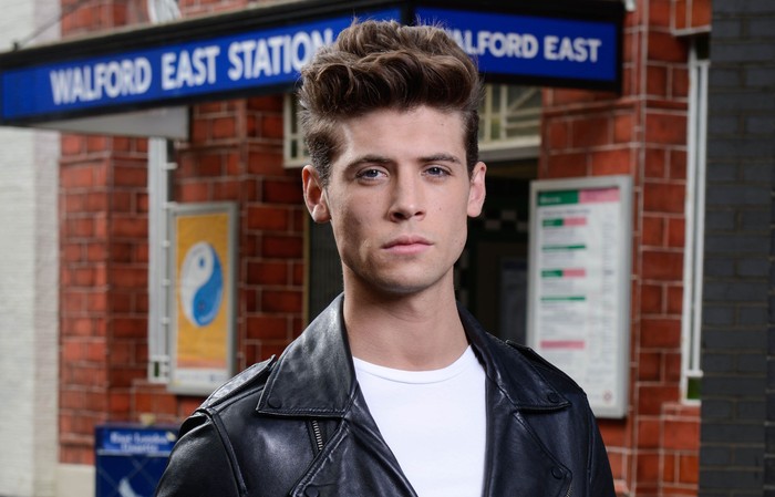 MarkFowlerJr Ned Porteous poses in a leather jacket and white t-shirt as Mark Fowler Jr outside Walford East Tube Station for EastEnders.