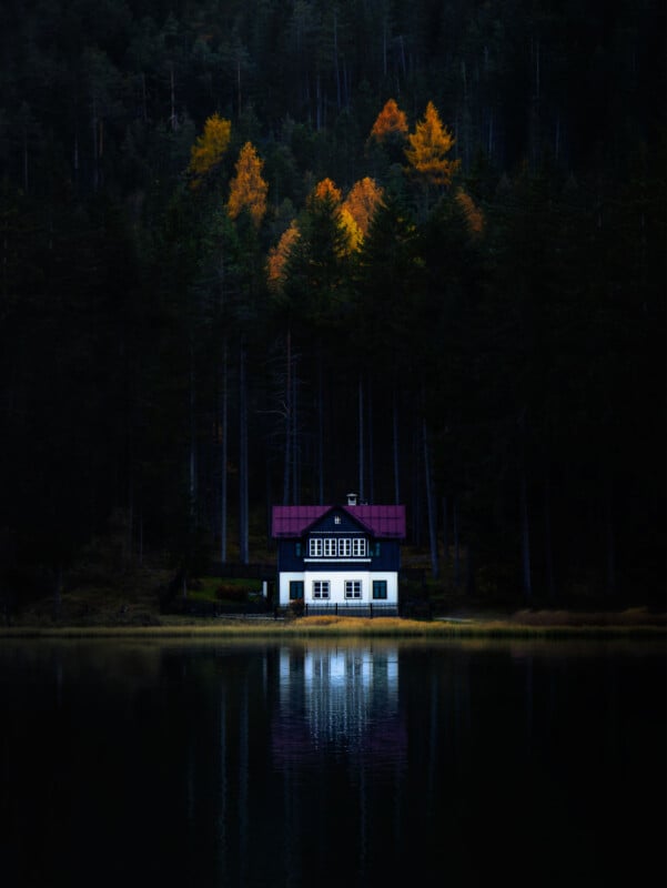A small white house with a red roof sits by a calm lake, surrounded by dark pine trees, with a few autumn-colored trees glowing in the background. The house and trees reflect in the still water.