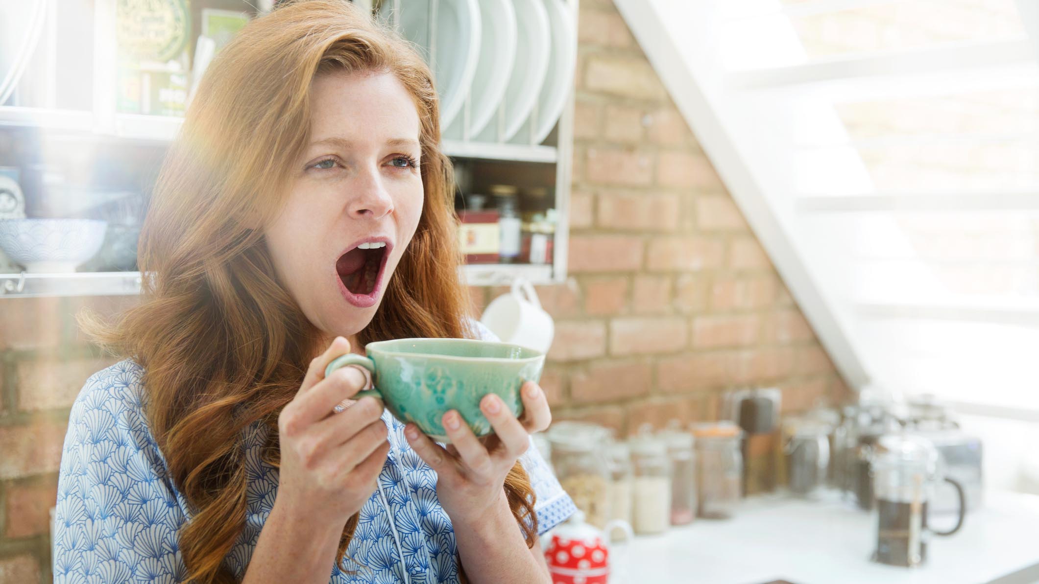 A woman with red hair yawns while drinking from a green cup