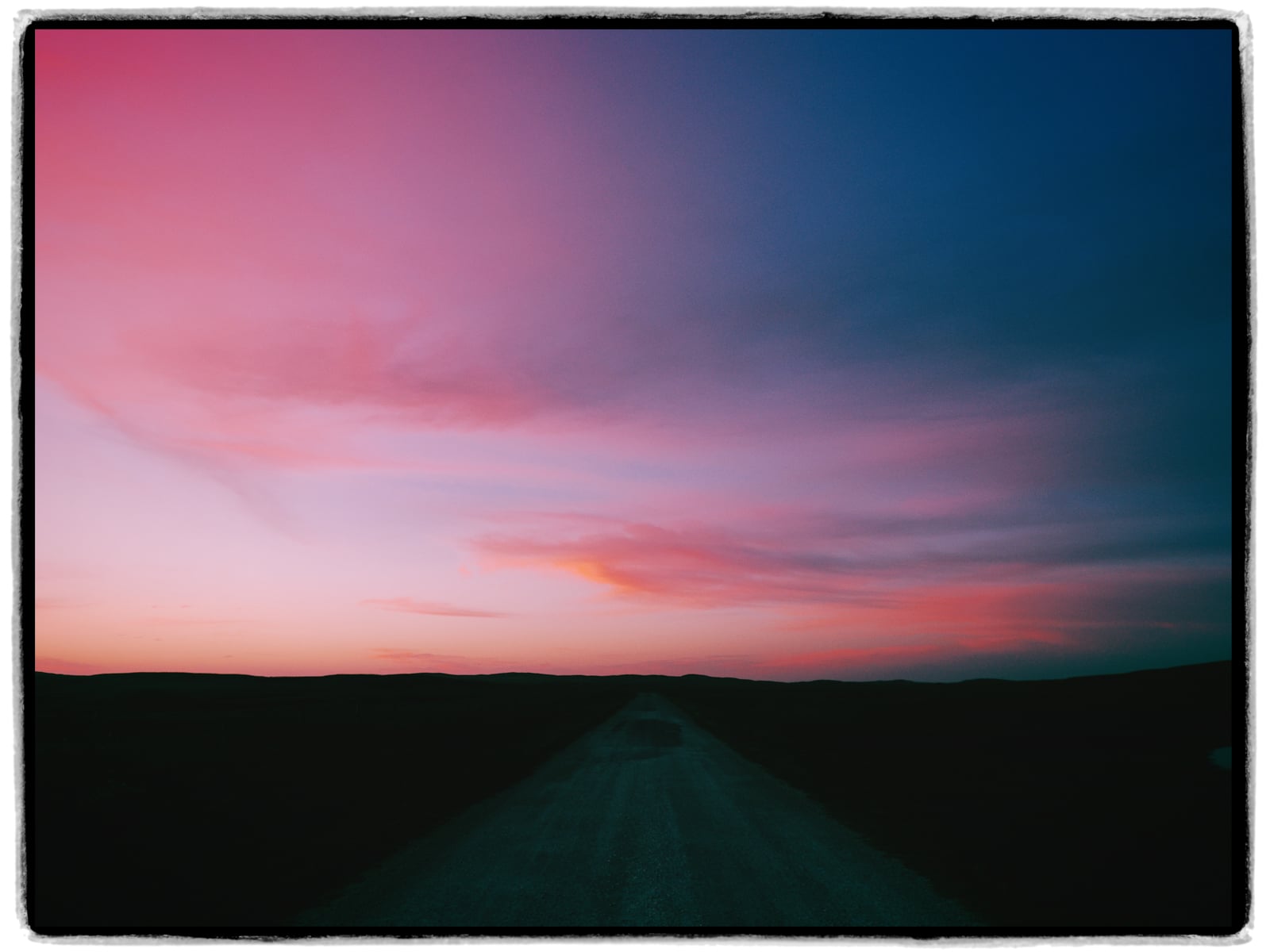 A deserted road stretches into the distance beneath a dramatic sky filled with pink, purple, and blue hues at sunset or sunrise. Dark silhouettes of hills line the horizon.