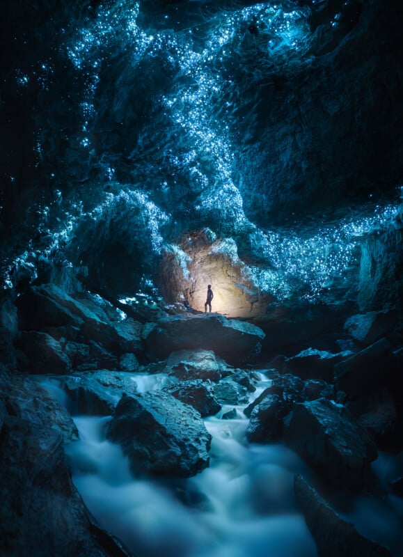 A person stands inside a rocky cave illuminated by glowing blue lights on the ceiling, with misty water flowing over rocks in the foreground.