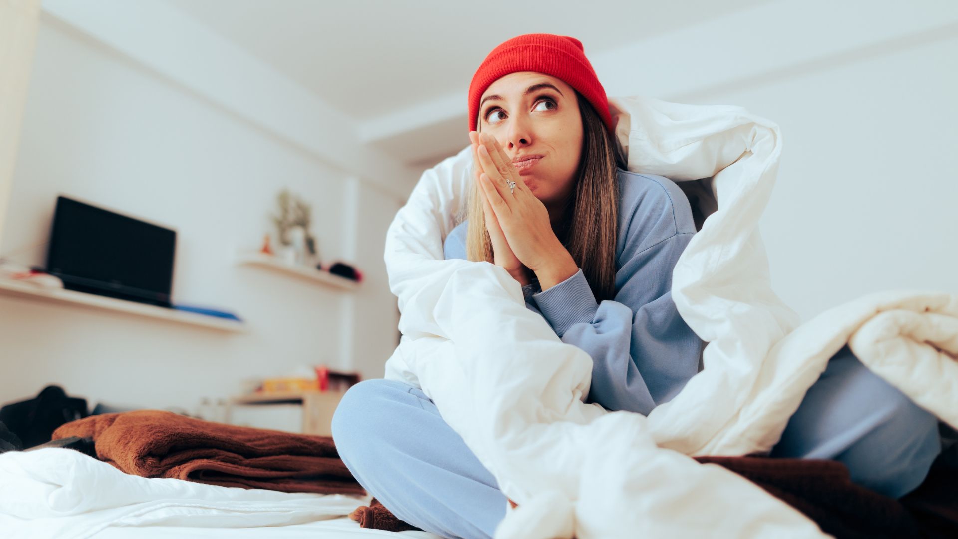 A woman sits up in bed wearing thick pajamas and a wooly hat. She is wrapped in a thick duvet.