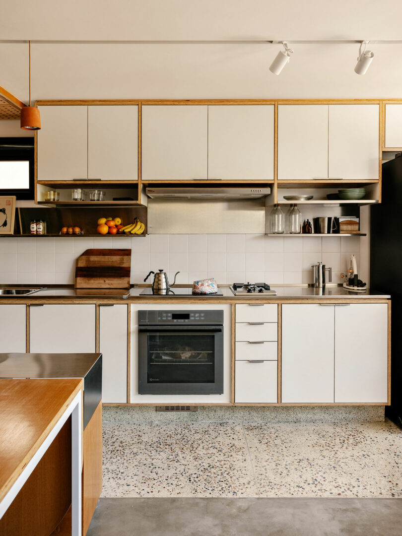 Modern kitchen with white cabinets, stainless steel appliances, a gas stove, oven, and open shelving displaying dishes, glassware, and fresh fruit.