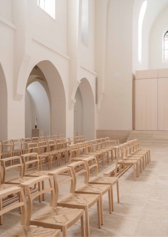 Rows of light wood chairs are arranged in a bright, minimalist church interior with white walls, arched doorways, high ceilings, and large windows letting in natural light.