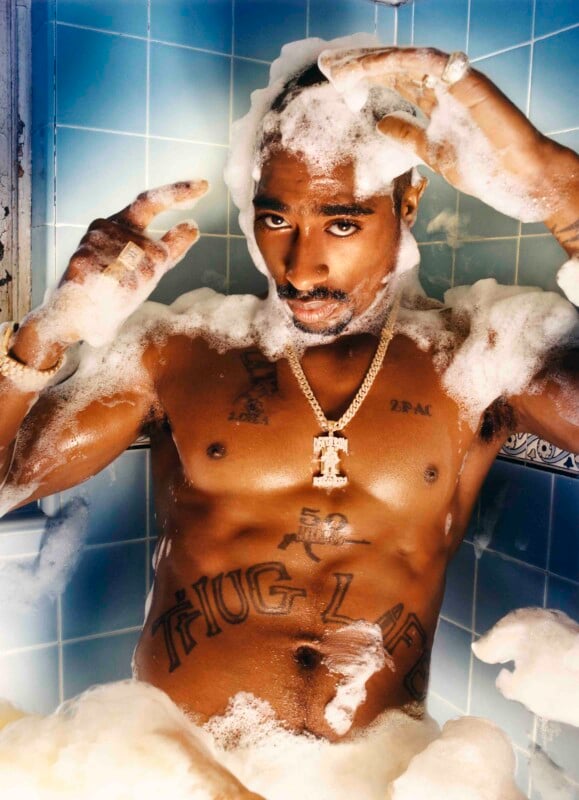 A shirtless man with tattoos relaxes in a bathtub filled with bubbles. He wears a gold chain and is surrounded by blue tiles, looking directly at the camera while raising his hands through the bubbles.